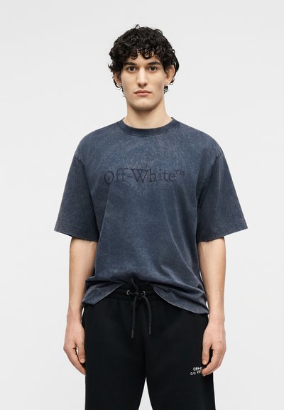 Νεαρός άνδρας με σγουρά μαλλιά, που φοράει μια ξεθωριασμένη σκούρη μπλε μπλούζα "Off-White" και μαύρο παντελόνι με κορδόνι, μπροστά σε απλό φόντο.