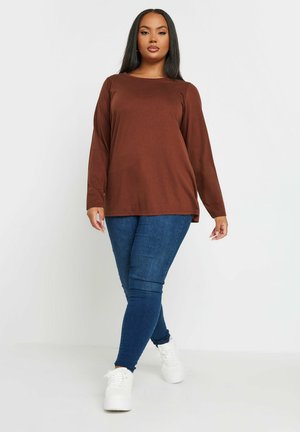 Bruine longslavenshirt in een relaxte pasvorm, gecombineerd met donkere skinny jeans en witte sneakers. Eenvoudig ontwerp zonder patronen of accenten.