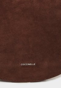 Borsa in suede marrone con una texture liscia, forma arrotondata e un logo argentato "COCCINELLE" in basso. Dettagli di cucitura visibili.