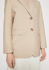 Μπεζ σακάκι με κομψή εφαρμογή, δύο καφέ κουμπιά και μπροστινές τσέπες. Λείο υλικό με γιακά τύπου notch και μακριά μανίκια.