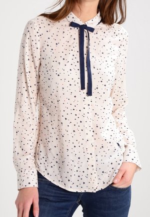 Femme portant un chemisier à manches longues de couleur claire avec de petites étoiles bleu marine et rouges, et un nœud papillon bleu marine au col, assorti à un jean foncé.