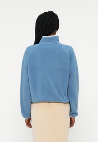 Pulover din fleece albastru cu guler înalt, mâneci raglan și o textură moale. Prezintă o tăietură scurtă și un design curat, minimalist.