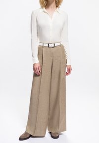 Femme portant une chemise blanche à boutons rentrée dans un pantalon beige taille haute à jambes larges, avec une ceinture blanche et des chaussures marron.