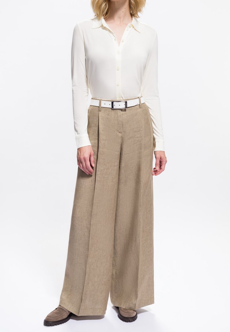 Femme portant une chemise blanche à boutons rentrée dans un pantalon beige taille haute à jambes larges, avec une ceinture blanche et des chaussures marron.