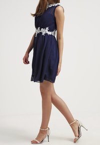Námořnická modrá bezramenná šaty s květinovým bílým krajkovým páskem, splývavý materiál a rozšířený střih. Nosí se s béžovými sandály na vysokém podpatku.