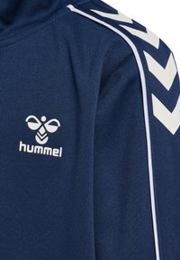 Marinblå sporttröja med hög krage, mjukt material, vita chevron-accenter på ärmarna och en tryckt logotyp framtill.
