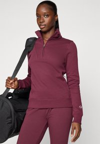 Burgunderfarbener Viertel-Reißverschluss-Sweatshirt mit gerippten Bündchen und Saum, kombiniert mit passenden Hosen. Das Modell hält eine schwarze Sporttasche.