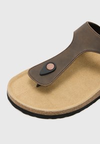 Brune læder flip-flop med en tekstureret beige ruskindssoled, der har en sort gummidetalje med en kobberknap og et minimalistisk design.