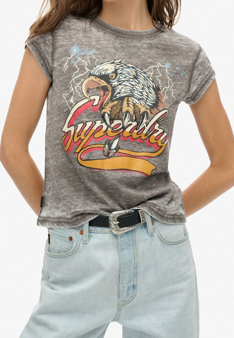 Šedé grafické tričko s designem orla, vysoce kontrastní červený a žlutý text "Superdry", krátké rukávy a volnější střih. Nahoře má na sobě světle modré džíny.