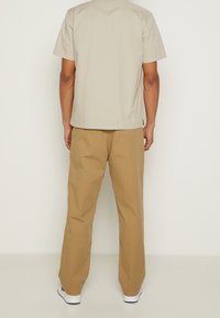 Beżowa koszula z krótkim rękawem oraz prostym dołem, w połączeniu z luźnymi spodniami w kolorze khaki. Buty są białe z czarnymi akcentami. Prosty design, materiał bawełniany.