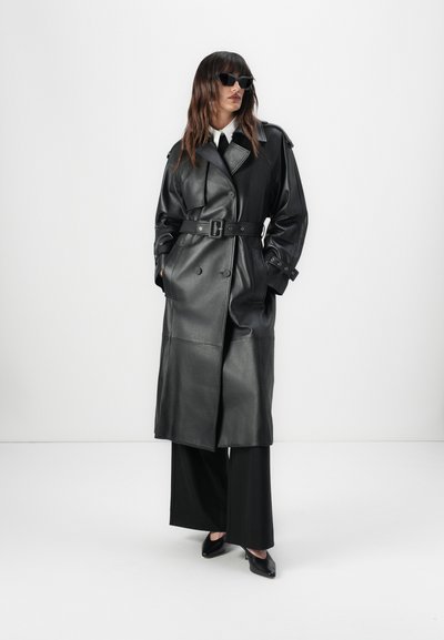 Long black leather trench coat with a belted waist, double-breasted design, and wide lapels. Model wears slim black trousers and sunglasses.
