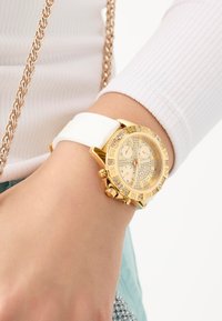 Montre dorée avec un bracelet en silicone blanc, accents en cristal et un cadran à motif présentant des sous-cadrans doubles et de grands index horaires.