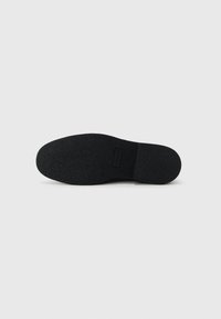 Suela de zapato de goma negra con superficie texturizada, bordes lisos y un parche de logo rectangular cerca del talón. Diseño simple y plano.