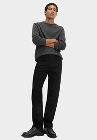 Pull en tricot gris, pantalon noir droit, et chaussures noires. Le modèle pose les bras croisés devant un fond gris clair.