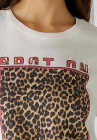 Bílý bavlněný tričko s designem leopardího vzoru a grafickým textem v červené a hnědé barvě nad vzorem. Kulatý výstřih, volný střih.