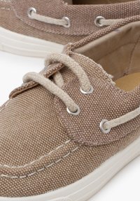 Braune Segelschuhe aus Canvas mit weißen Gummisohlen. Merkmale sind beige Schnürsenkel, Metallösen und strukturierter Stoff.