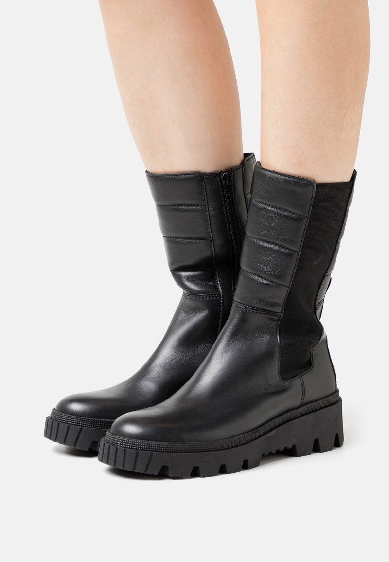 Bottes en cuir noires mi-mollet avec semelles épaisses à rainures et fermetures éclair latérales, portées sur des jambes nues sur un fond blanc.