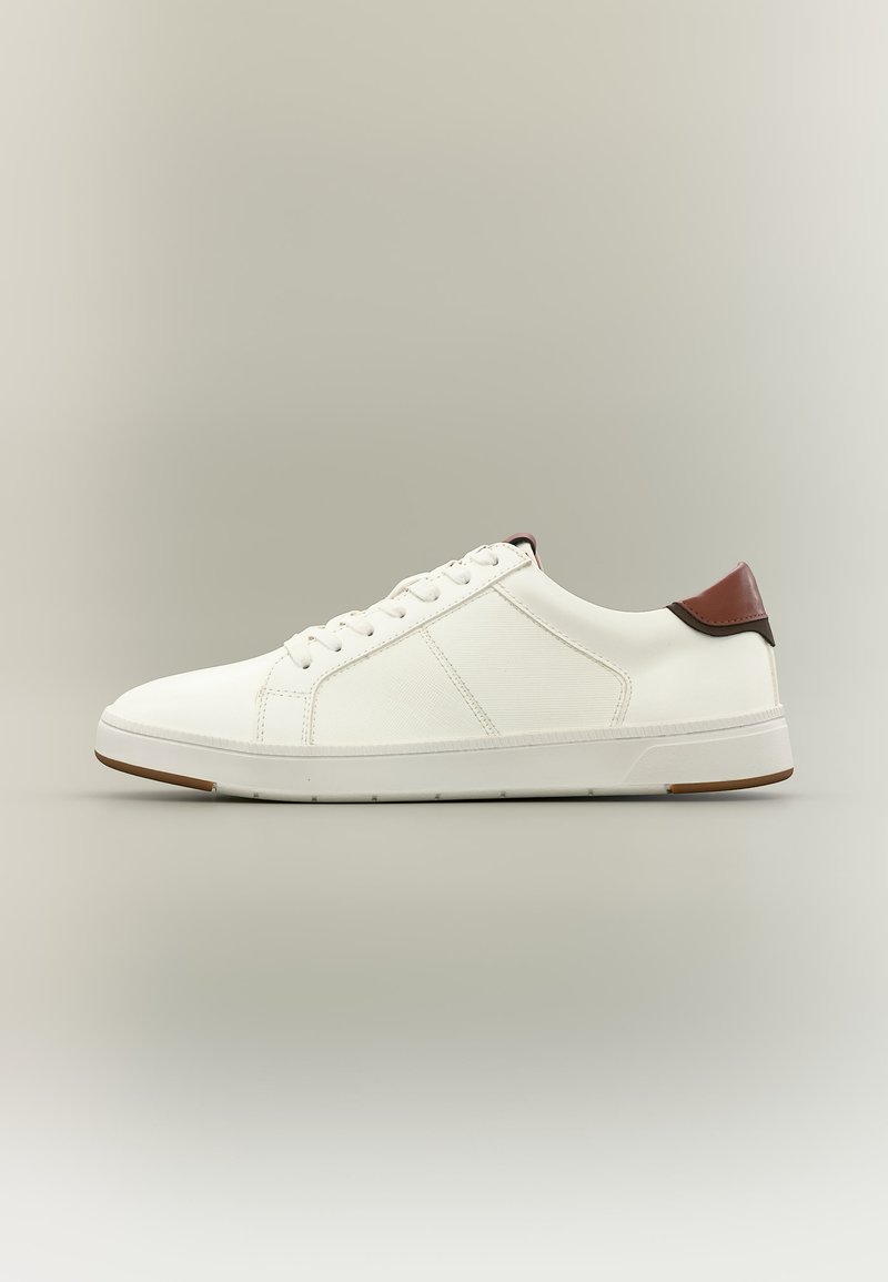Baskets basses blanches avec lacets blancs, panneaux latéraux texturés et languette de talon marron sur un fond neutre.