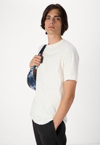 Hvid kortærmet t-shirt med rund hals. Modellen holder en blå tie-dye taske med en sort skulderrem mod mørke bukser.