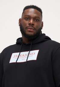 Black hoodie with a front graphic label in blue and red text. Features a kangaroo pocket and drawstrings on the hood. Soft fabric texture.