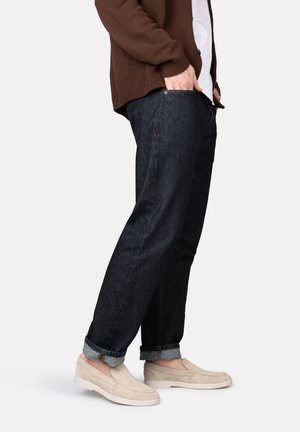 Uomo con jeans scuri risvoltati, scarpe beige slip-on, maglione marrone e camicia bianca, in piedi con una mano in tasca su sfondo bianco.