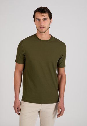 Homme aux cheveux courts et foncés, portant un t-shirt à manches courtes vert olive et un pantalon beige, debout devant un fond clair uni.