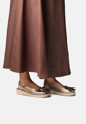 Fødder iført metalliske gyldne espadrilles med sløjfedetalje og ankelstropper, kombineret med en brun, flydende midi-nederdel.