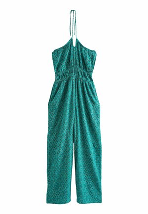 Femme portant une combinaison dos nu à motif turquoise et des sandales compensées noires, debout à l'extérieur près d'assises blanches rembourrées.