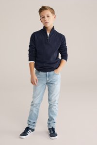 Tumši zils džemperis ar ceturtdaļzipu, kopā ar gaiši zilām džinsu biksēm. Apģērbu papildina tumši zili sporta apavi ar baltām svītrām un fakturētu audumu.