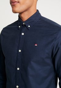 Námořnicky modrá košile na knoflíky se standardním límcem, s bílými knoflíky a malým červeným logem hory na levé části hrudi. Hladký povrch.