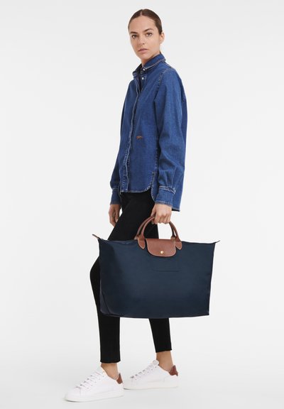 Navy nylon tote bag with brown leather accents and handles, featuring a top zip, spacious design, and a logo plate on the front.