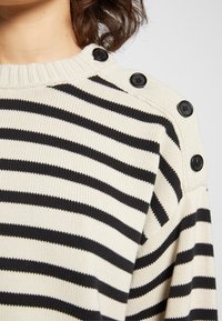 Gros plan d'un pull en maille rayé noir et blanc avec des boutons noirs le long de la couture de l'épaule et un col côtelé.