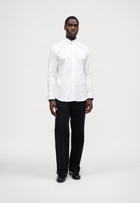 Camisa branca de manga comprida com botões e gola alta, combinada com calças pretas e sapatos pretos. Design simples e limpo, sem padrões.