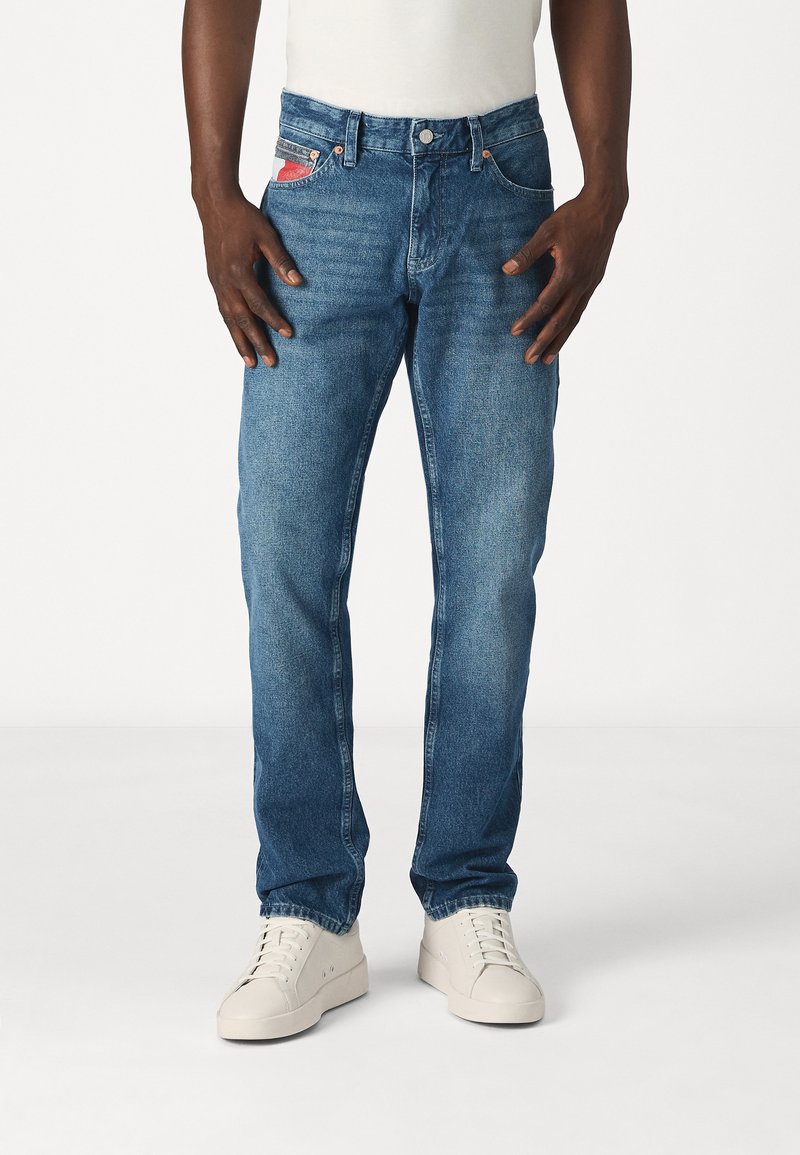 Jeans de mezclilla azul con un corte recto, que presentan un ligero desvanecimiento, cinco bolsillos y detalles de costura en naranja. Llevados con zapatillas blancas.