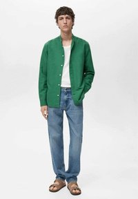 Green button-up shirt over a white t-shirt, paired with light blue jeans and brown sandals; the shirt features a collarless design.