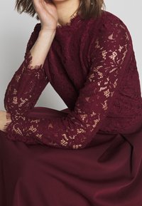 Top en dentelle bordeaux à manches longues présentant des motifs floraux complexes, des bords festonnés et un corps en tissu lisse.