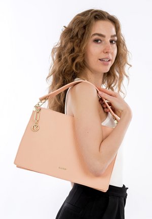 Frau mit lockigem Haar, die eine große beigefarbene Lederhandtasche über der Schulter trägt, bekleidet mit einem weißen Tanktop und schwarzen Hosen.