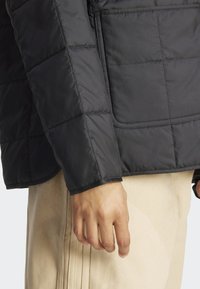 Gros plan sur une main reposant à côté de pantalons beiges avec des coutures visibles, portant une veste matelassée noire avec des coutures en motif carré.