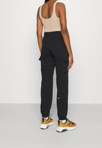 Černé cargo tepláky s dvěma bočními kapsami, mírným manžetovým zakončením u kotníků, skombinované s vícebarevnými teniskami a béžovým tílkem.