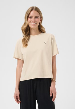 Femme aux longs cheveux blonds ondulés portant un t-shirt beige à manches courtes avec un petit cœur noir sur la poitrine et un pantalon noir ample, souriante.