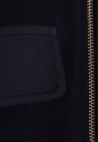 Giacca di tessuto blu navy con tasca a toppa testurizzata e zip in metallo dorato, caratterizzata da una finitura liscia e dettagli di cucitura distintivi.