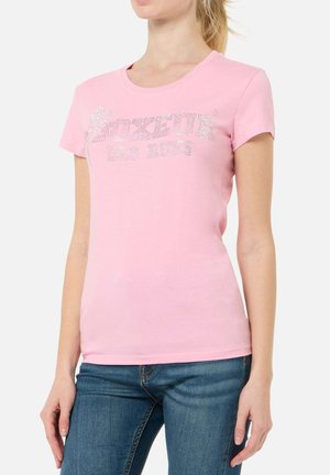 Donna che indossa una t-shirt rosa a maniche corte con scritta argento e jeans blu scuro, in piedi davanti a uno sfondo bianco semplice.