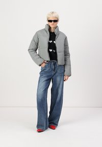 Giacca puffer grigia chiara, design cropped, abbinata a un maglione nero con motivi a cuore bianchi e jeans a vita alta blu. Scarpe rosse.