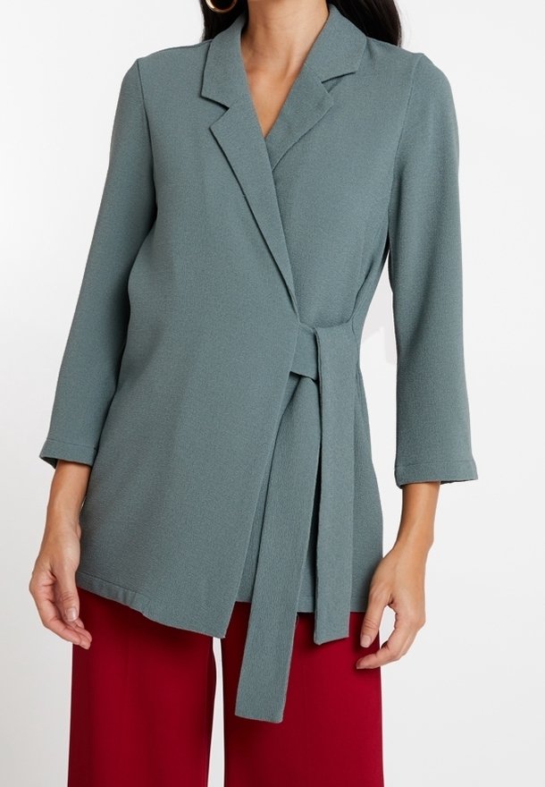 Světle tyrkysový kabát s texturovanou tkaninou, s klopami, tříčtvrtečními rukávy a detailem zavázaného pásku, v kombinaci s červenými kalhotami.