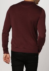 Donkerrode trui met lange mouwen, een ronde halslijn en geribbelde zoom. Gladde textuur en een aansluitend ontwerp, gedragen met zwarte broek.