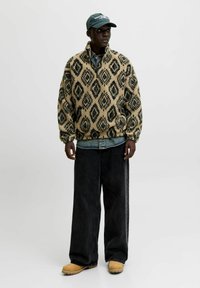 Beiger Fleecepullover mit schwarzem Diamantmuster; verfügt über einen Halbreißverschluss-Kragen, lockere Passform, getragen über einem Jeanshemd und kombiniert mit weiten schwarzen Jeans.