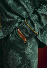 Zelený textil s kvetinovým vzorom a zlatými lemami. Obsahuje nastaviteľný manžet a integrovanú kapsu, textúrovaný povrch.