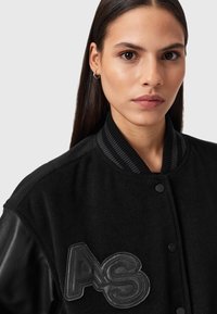 Giovane donna con capelli scuri e lisci, che indossa una giacca nera con maniche in pelle e un patch ricamato con "AS" sul petto, su uno sfondo neutro.