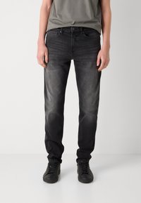 Jeans de mezclilla negra con un acabado desgastado, ajuste Slim, cinco bolsillos y cierre de botón. Combinados con zapatillas negras y una camiseta gris.