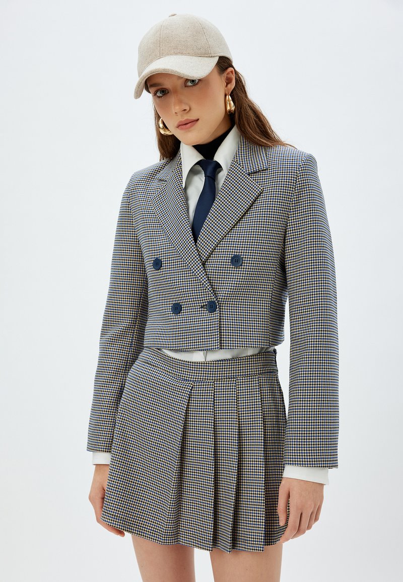 Traje de cuadros con chaqueta corta de doble botonadura y falda plisada. Incluye una gorra de color claro y una corbata azul marino.